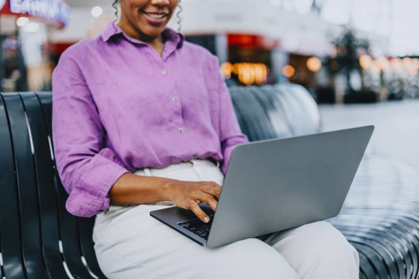 A woman in a purple shirt is using a laptop on a bench in a shopping mall, smiling and engaged. The atmosphere is relaxed and productive.